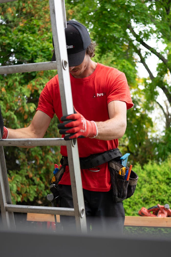A gardener wearing safety gear climbs a ladder outdoors, surrounded by green foliage.